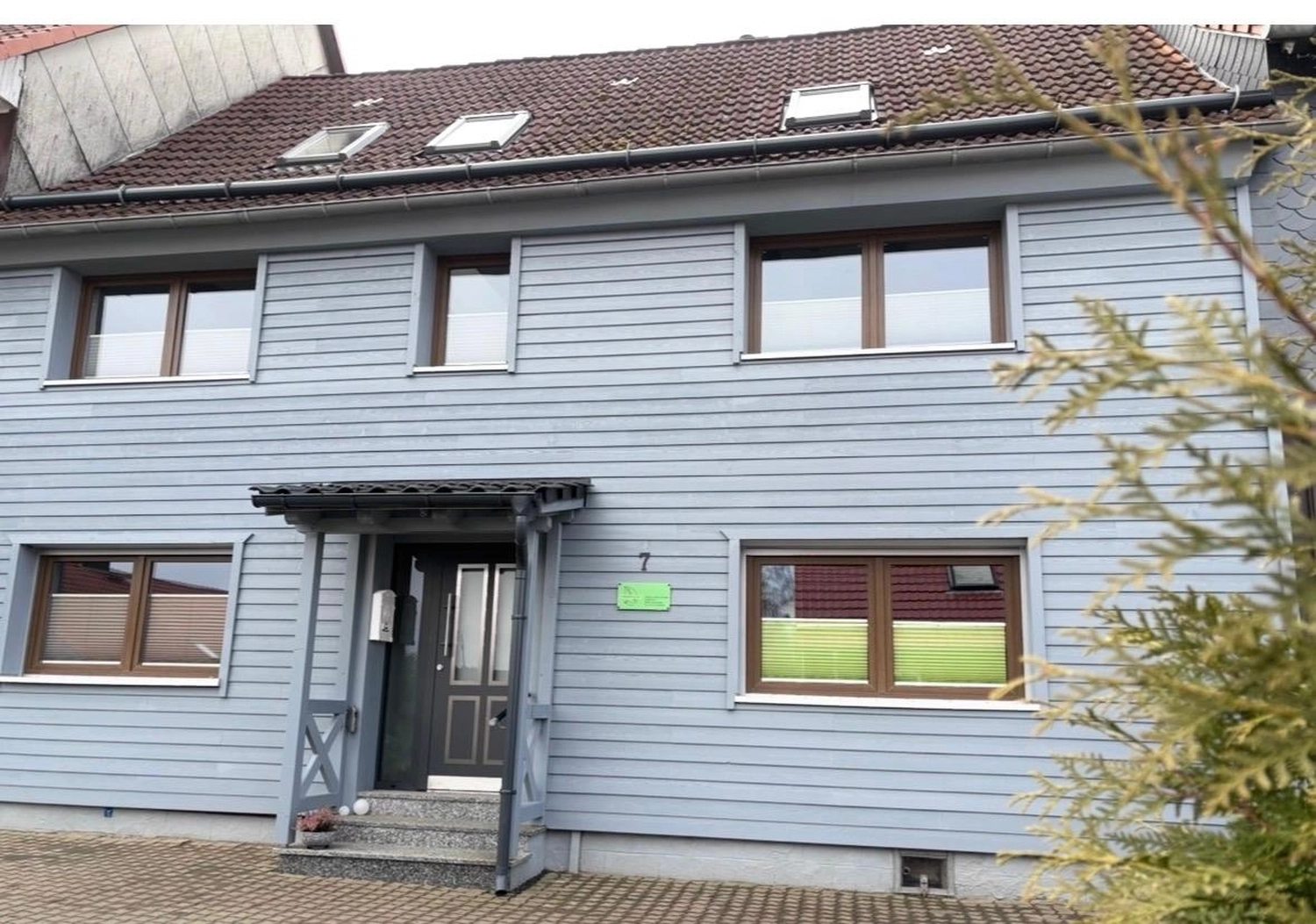 Ecklebe - Ferienwohnung - Die Vorderansicht eines zweistöckigen Hauses mit hellblaugrauer Verkleidung und braun gerahmten Fenstern.