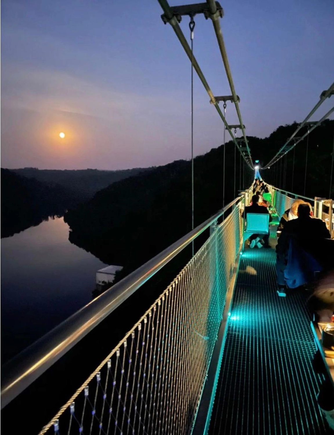 Ecklebe - Ferienwohnung - Eine Nacht auf einer Hängebrücke aus Metall, mit Blick auf ein Gewässer und dem Mond am Himmel.