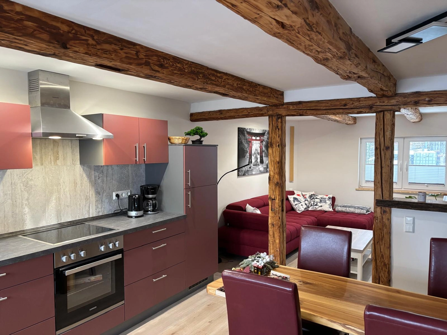 Ecklebe - Ferienwohnung - Eine moderne Küche mit bordeauxroten Schränken und Edelstahlgeräten, die sich zu einem Wohnbereich mit rotem Sofa und Holzbalken an der Decke öffnet.