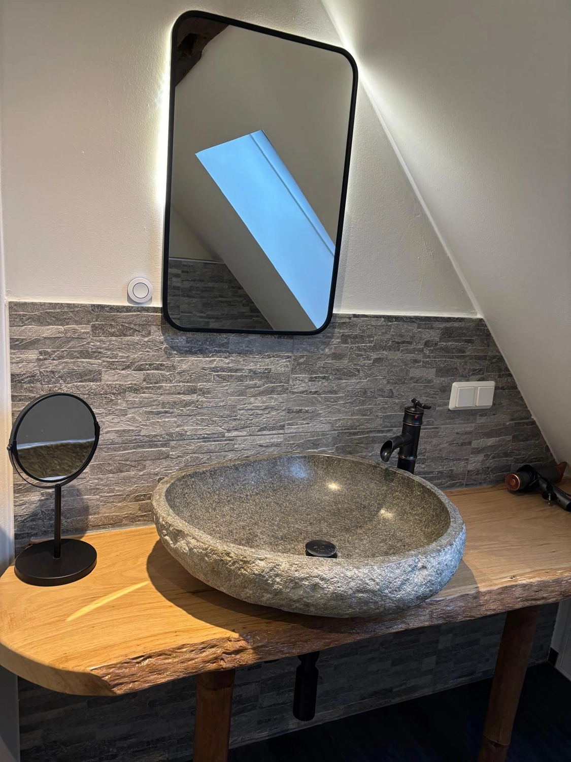 Ecklebe - Ferienwohnung - Blick auf den Waschbeckenbereich eines Badezimmers mit einem einzigartigen, runden Steinbecken, einem darüber hängenden Spiegel und grauen, steingefliesten Wänden.