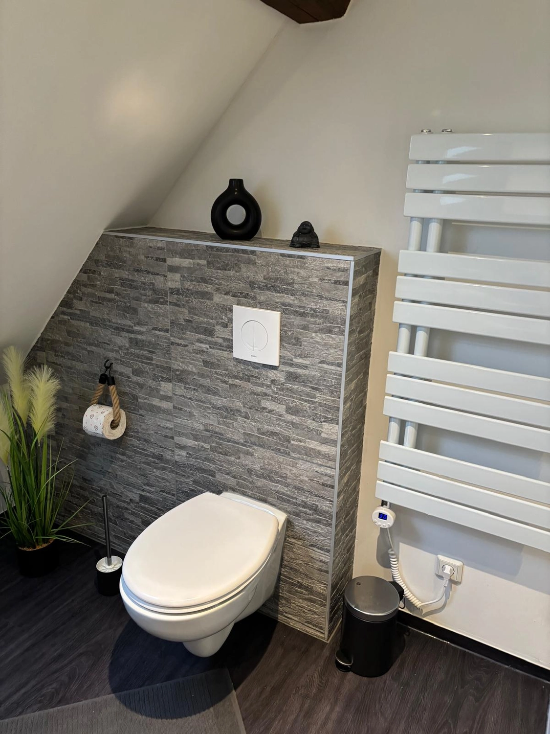 Ecklebe - Ferienwohnung - Ein Badezimmer mit Toilette, dahinter eine graue, steingeflieste Wand und ein kleines Regal mit einem Dekorationsgegenstand.