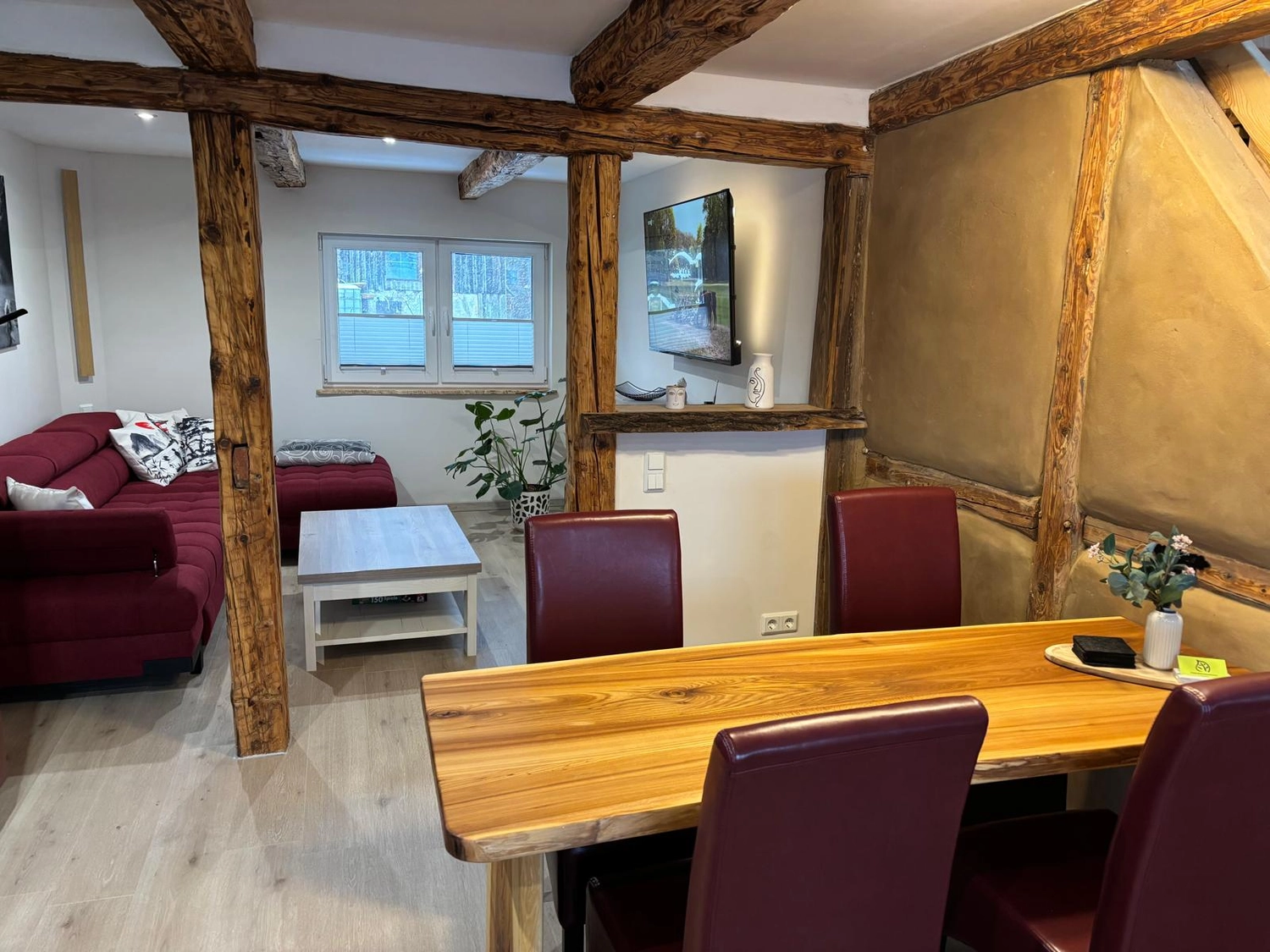 Ecklebe - Ferienwohnung - Ein Essbereich mit freiliegenden Holzbalken, einem hölzernen Esstisch, roten Stühlen und einer kleinen Sitzecke im Hintergrund.