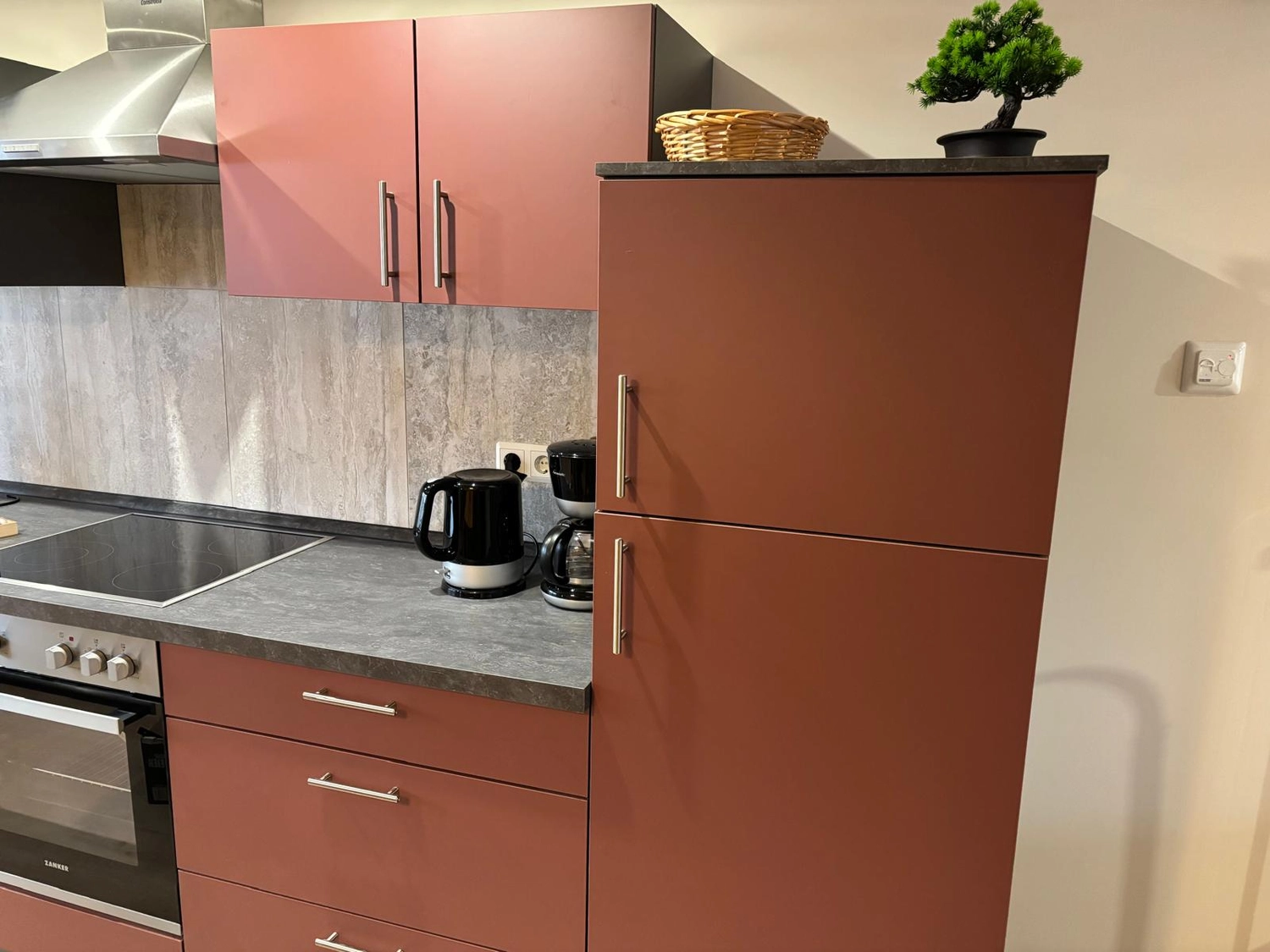 Ecklebe - Ferienwohnung - Eine Küche mit terrakottafarbenen Schränken, einer dunkelgrauen Arbeitsplatte und einem kleinen Bonsai-Baum auf der Arbeitsplatte.