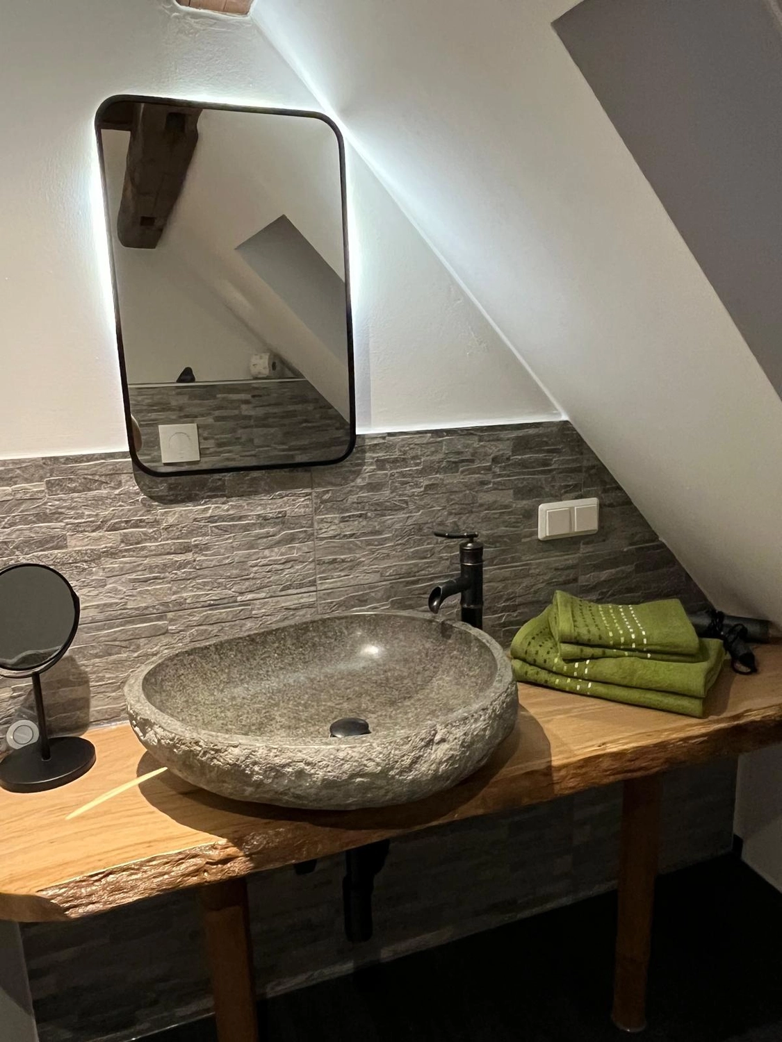 Ecklebe - Ferienwohnung - Ein modernes Badezimmer mit einem einzigartigen Steinwaschbecken auf einer Holzablage, einem großen Spiegel und grünen Handtüchern.