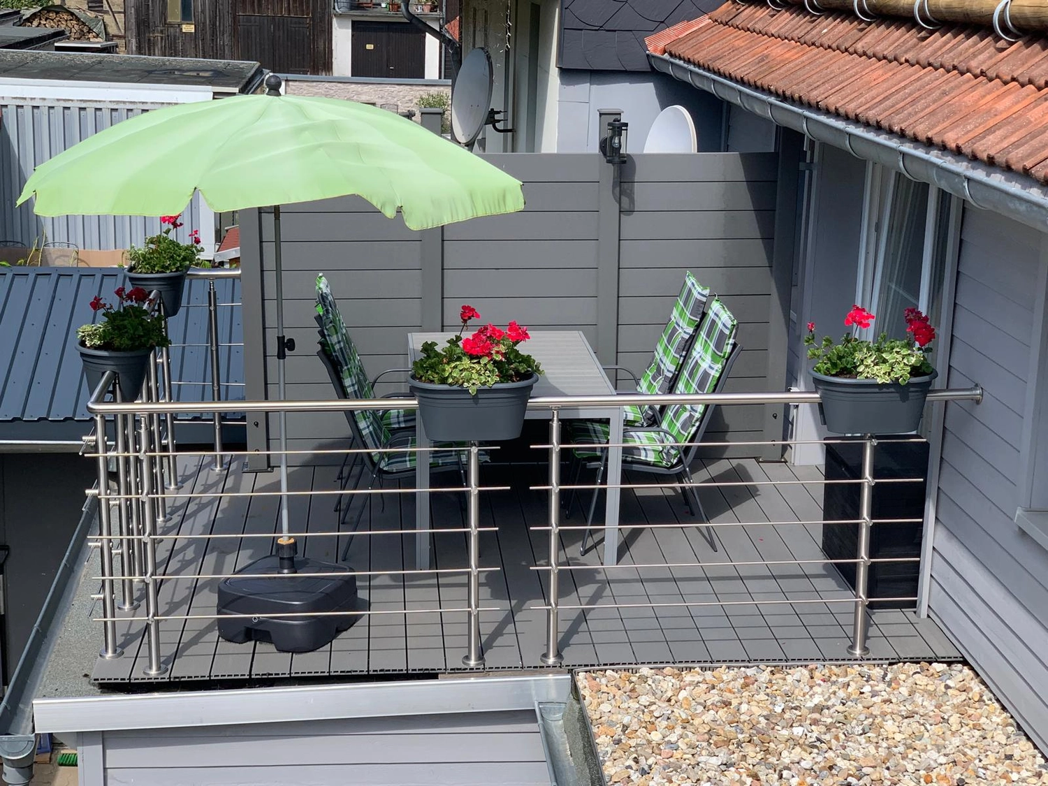 Ecklebe - Ferienwohnung - Ein sommerlicher Blick auf dieselbe Terrasse mit einem grünen Sonnenschirm, roten Topfblumen und einer grauen Holzfassade.