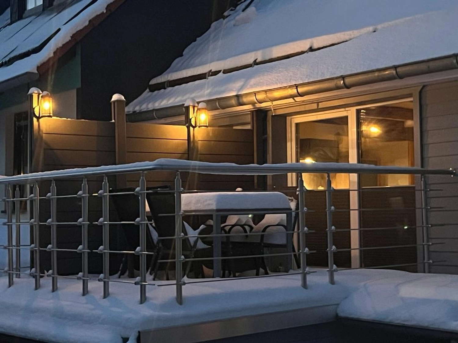 Ecklebe - Ferienwohnung - Eine schneebedeckte Terrasse mit Edelstahlgeländer, Tisch und Stühlen sowie warmen Lichtern an der Seite eines Holzgebäudes.