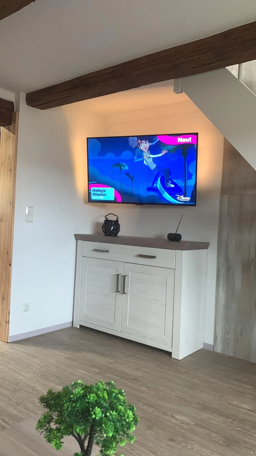 Ecklebe - Ferienwohnung - Über einem weißen zweitürigen Schrank hängt ein an der Wand montierter Fernseher, auf dem ein farbenfrohes Videospiel oder ein Musikvideo läuft.