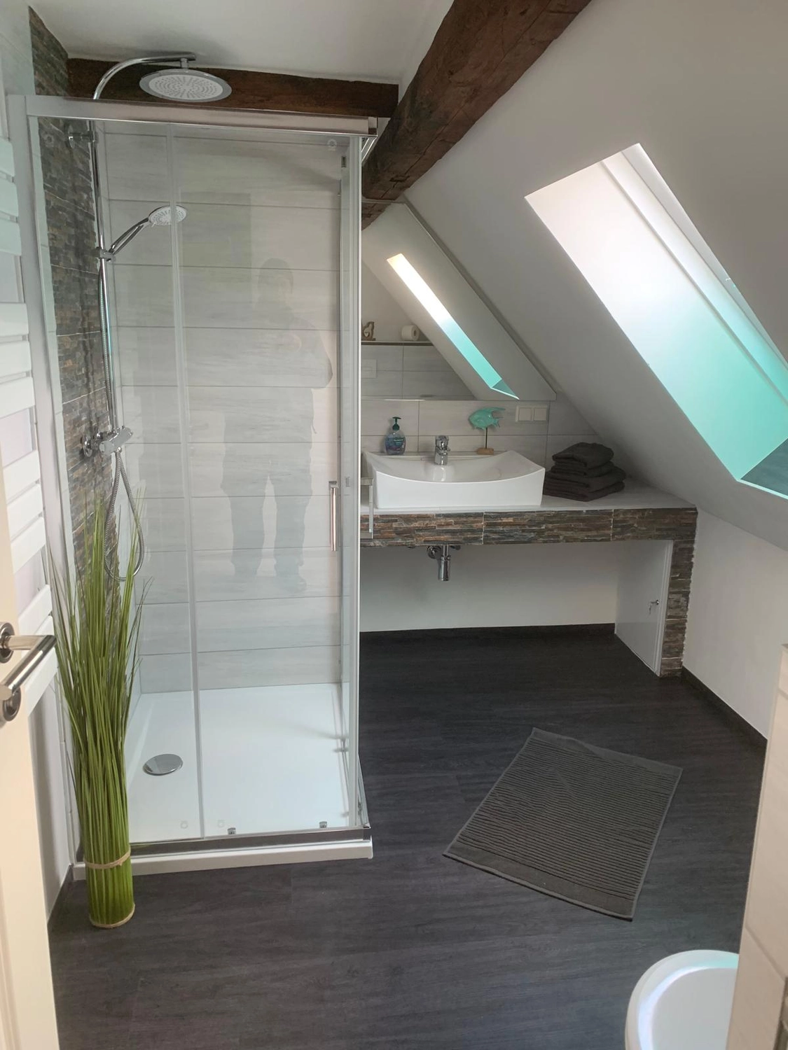 Ecklebe - Ferienwohnung - Eine moderne, ebenerdige Dusche mit weißen Fliesen und Glastür sowie einem weißen Waschbecken auf einem Holzgestell unter einer schrägen Decke mit Oberlicht.