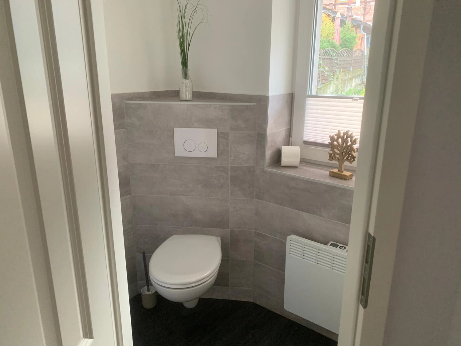 Ecklebe - Ferienwohnung - Ein kleines Badezimmer mit Toilette, grau gefliesten Wänden und einem Fenster mit Blick auf ein Haus.