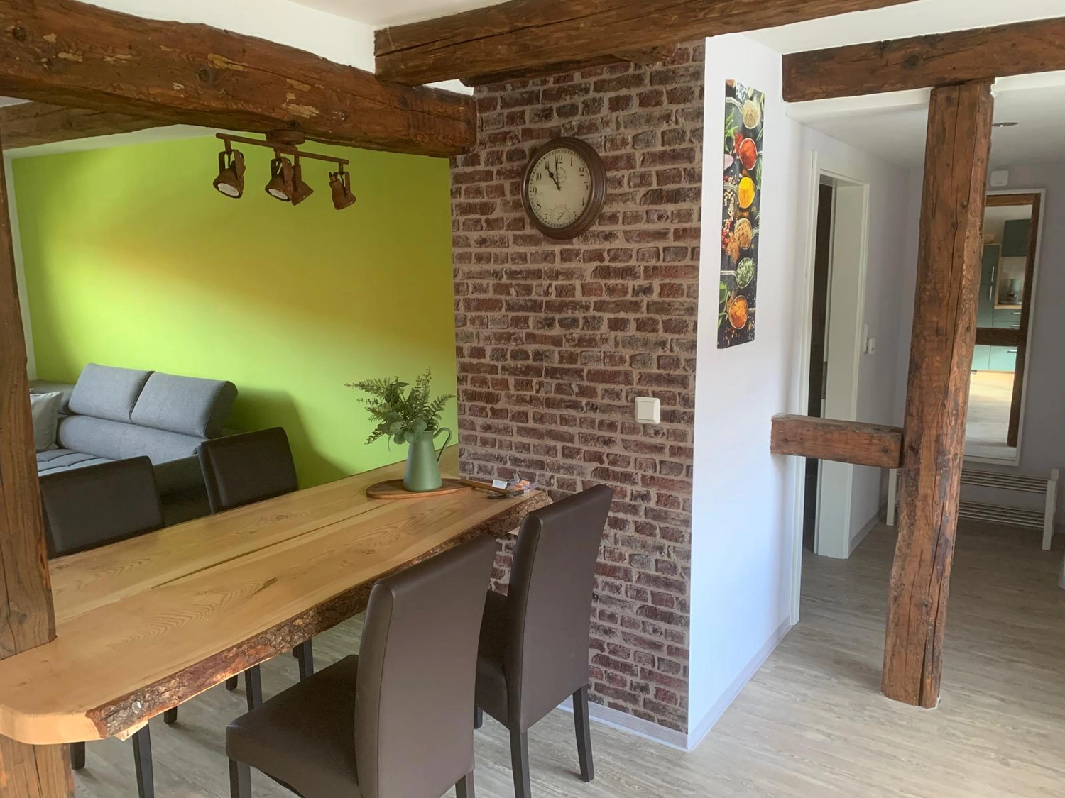 Ecklebe - Ferienwohnung - Ein Essbereich mit einem langen Holztisch und Stühlen grenzt an einen Wohnbereich mit einer grünen Akzentwand und einem Sofa.