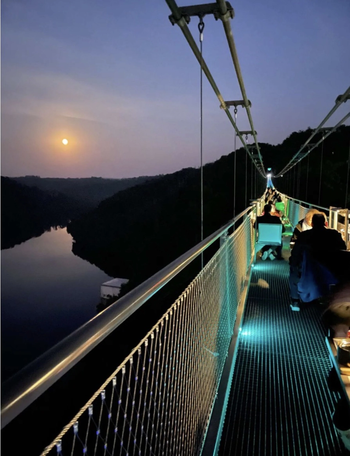 Ecklebe - Ferienwohnung - Eine Nacht auf einer Hängebrücke aus Metall, mit Blick auf ein Gewässer und dem Mond am Himmel.