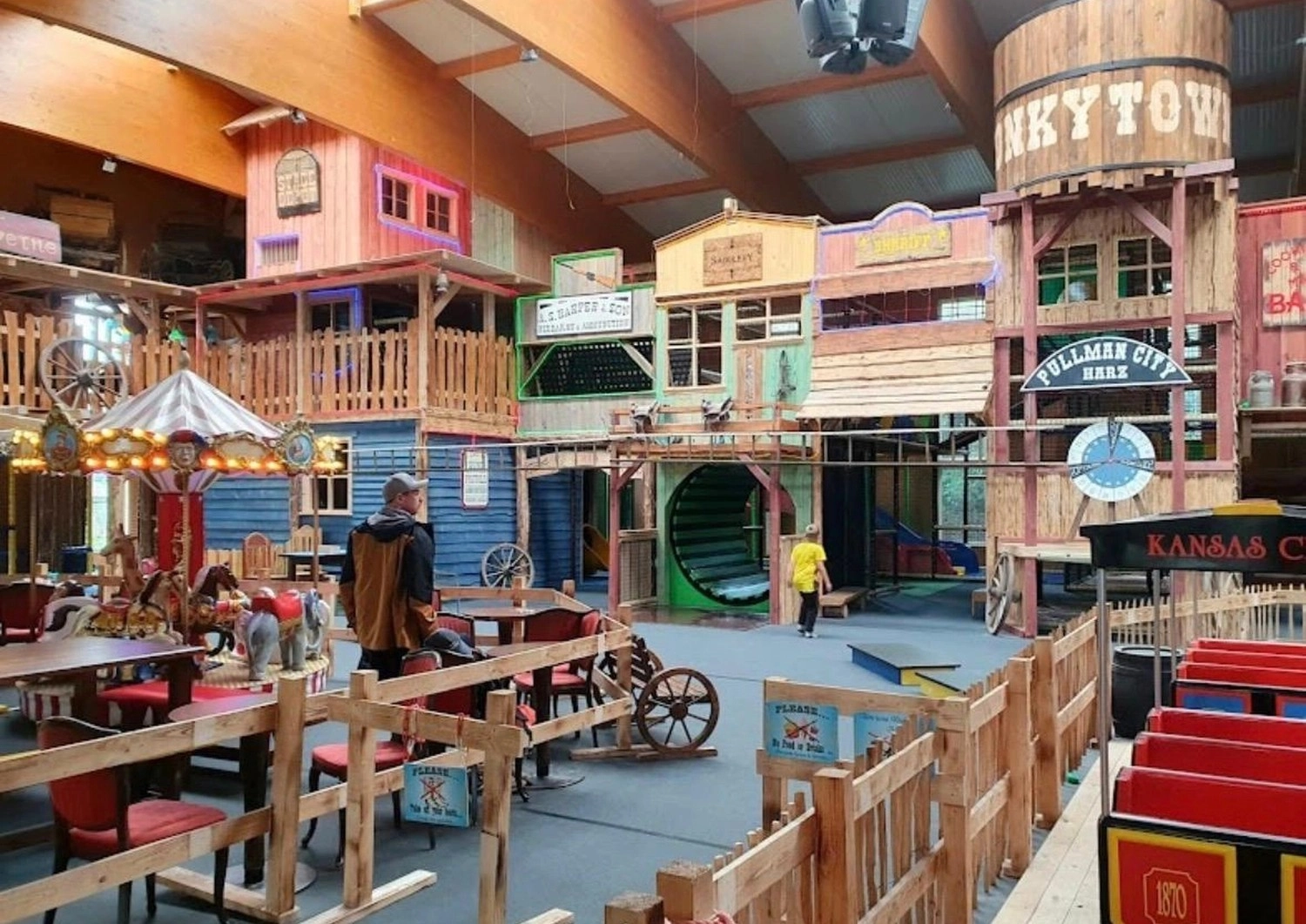 Ecklebe - Ferienwohnung - Ein Indoor-Spielbereich, der wie eine mehrstöckige Westernstadt gestaltet ist und verschiedene Bauwerke, ein kleines Karussell und Holztische umfasst.